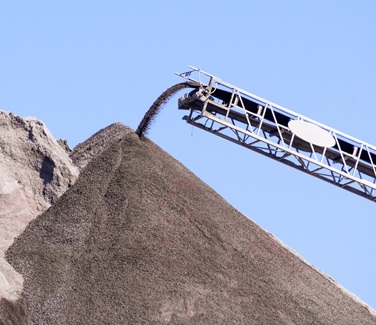 Ein Förderband transportiert feinen Kies oder Sand und lässt das Material an seinem Ende auf einen großen, kegelförmigen Schotterhaufen fallen. Der Himmel im Hintergrund ist klar und blau.

                          